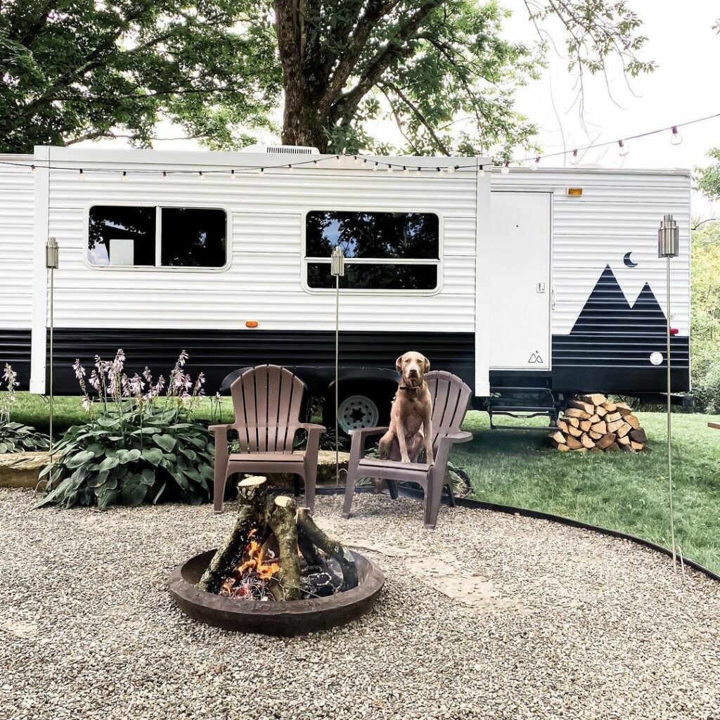 Keystone Travel Trailer RV parked with a firepit and landscaping in the forefront. A brown dog sites in one of two chairs staring at the camera. 