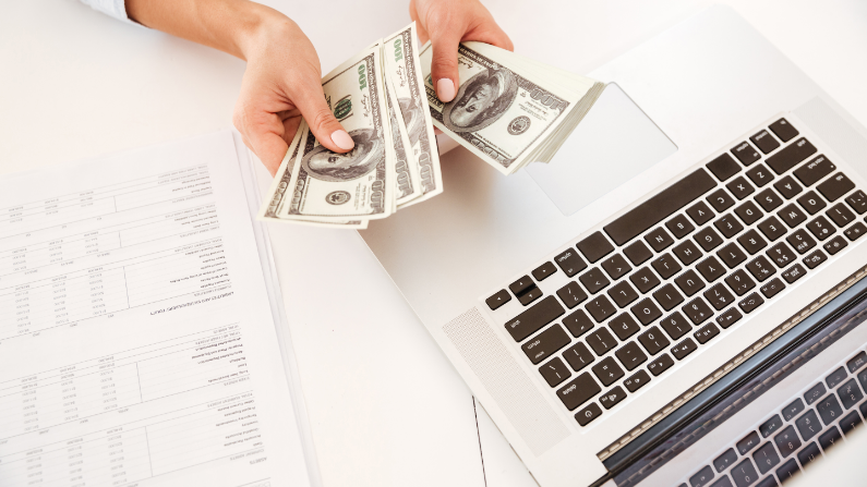 hands counting cash with a laptop and white desk in the background