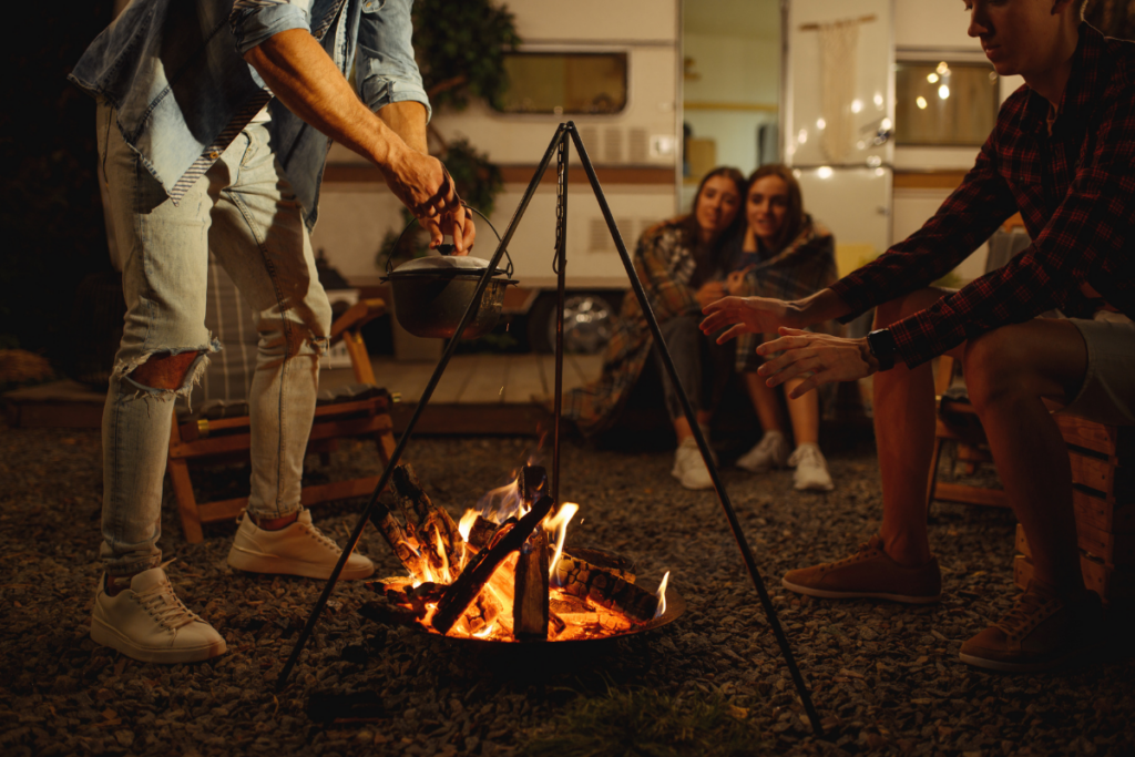 Group of 4 friends sitting around a campfire with an RV in the background